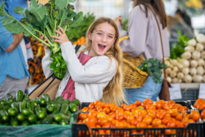 Young girl smiling excitedly while holding a large bunch of leafy greens at a farmers market stall, surrounded by fresh tomatoes and cucumbers.