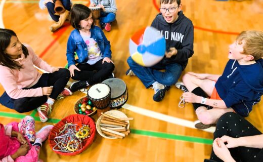Children sitting in a circle playing music and rhythm games with drums, shakers, and a beach ball.