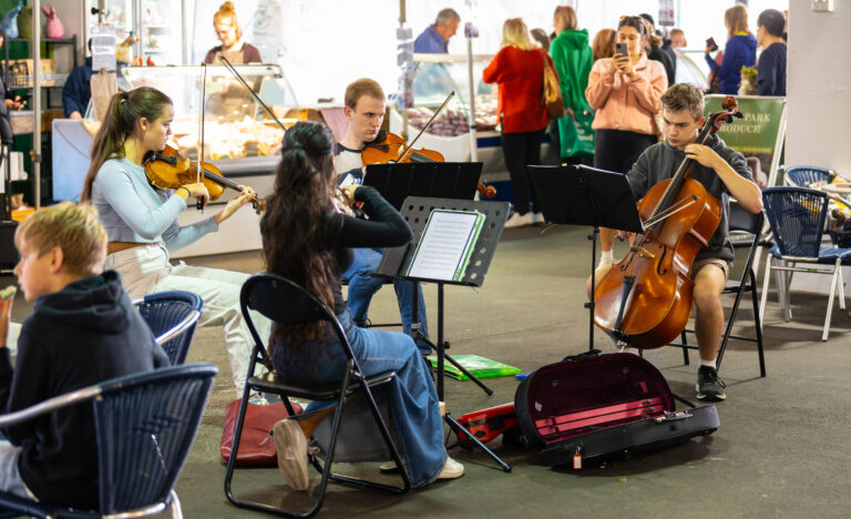 Four young musicians perform as a string quartet inside the Adelaide Showground Farmers’ Market, surrounded by marketgoers enjoying the music.