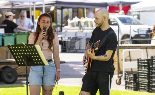 Ebony sings into a microphone while Dylan plays guitar at the Adelaide Showground Farmers’ Market, performing outdoors under the market tents.