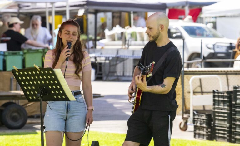 Ebony sings into a microphone while Dylan plays guitar at the Adelaide Showground Farmers’ Market, performing outdoors under the market tents.