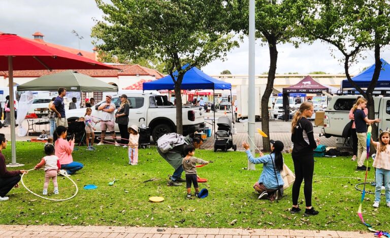 Children and families enjoy circus activities on the grass under red umbrellas at the Adelaide Showground Farmers’ Market during a Come and Try session with Lolly Jar Circus.