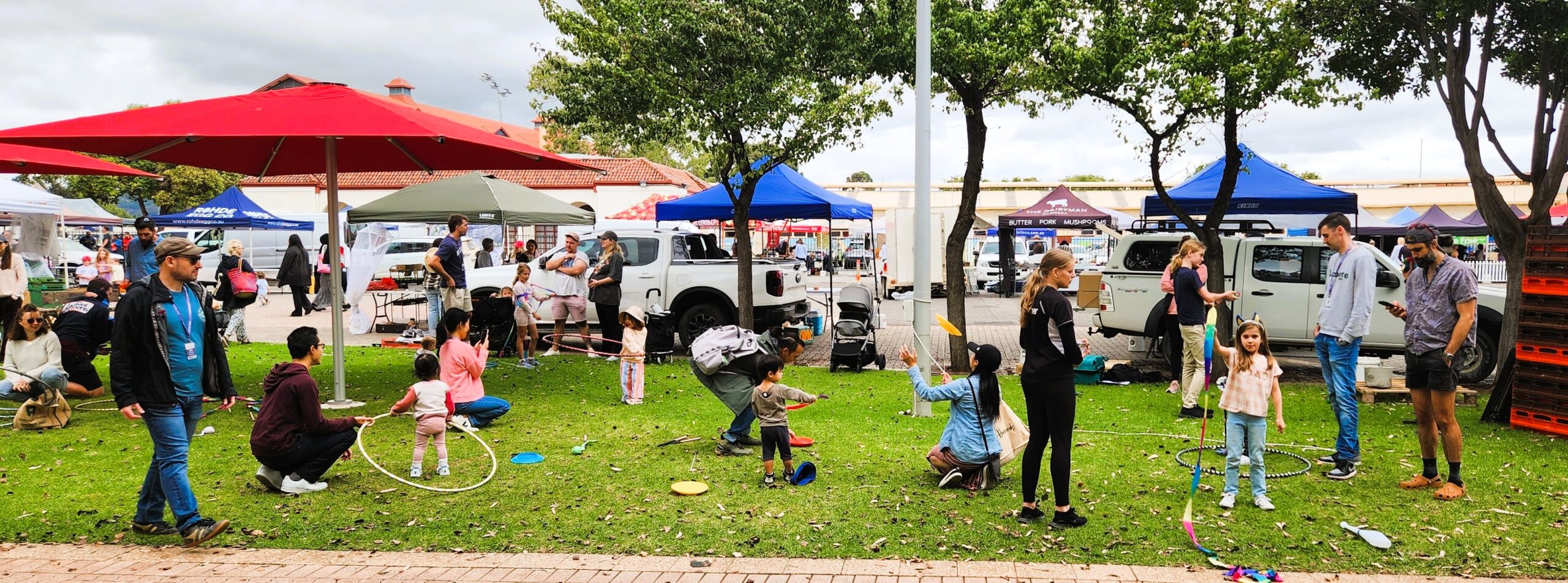 Children and families enjoy circus activities on the grass under red umbrellas at the Adelaide Showground Farmers’ Market during a Come and Try session with Lolly Jar Circus.