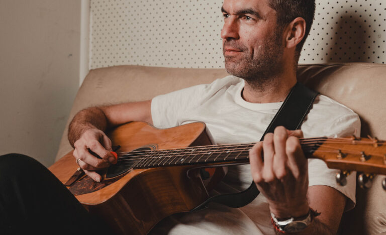 A man sitting on a couch playing an acoustic guitar, looking ahead with a relaxed, focused expression.