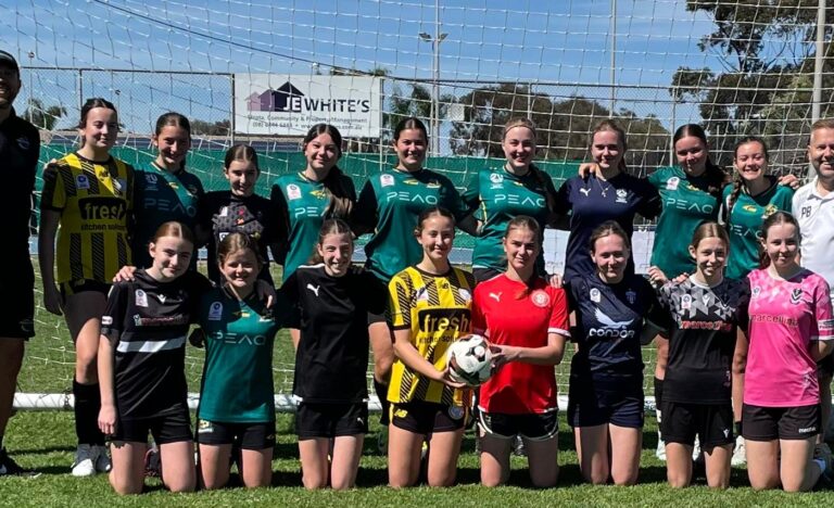 Group photo of Believe & Achieve junior girls soccer players and coaches standing in front of a goal on a sunny day.