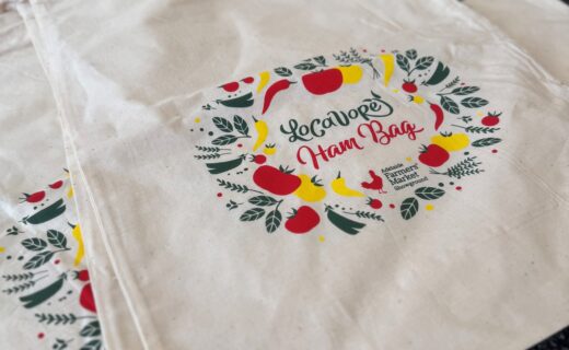 A stack of cream-coloured reusable ham bags with a colourful wreath design featuring fruit and leaves and the words “Locavore Ham Bag” printed on the front.