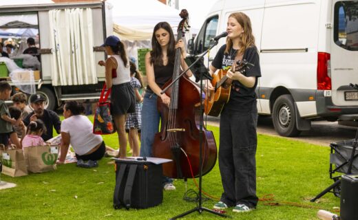 Two young musicians perform outdoors at the Adelaide Showground Farmers’ Market, one playing guitar and singing, the other on double bass, with marketgoers relaxing nearby on the grass.