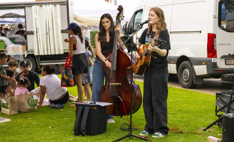 Two young musicians perform outdoors at the Adelaide Showground Farmers’ Market, one playing guitar and singing, the other on double bass, with marketgoers relaxing nearby on the grass.