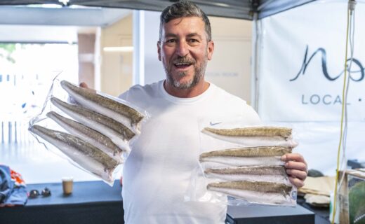 A stallholder at The Local Fish stands inside the market pavilion smiling and holding two large South Australian lobsters wrapped in netting.