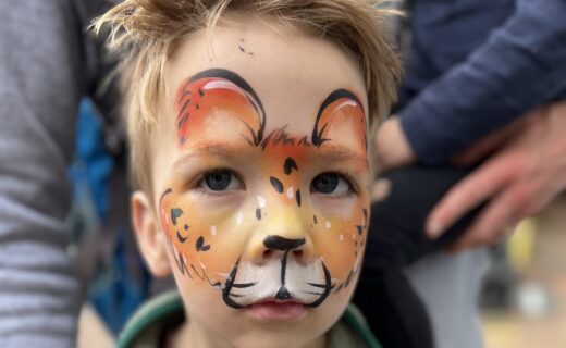 Young child with detailed lion face paint in warm orange and brown tones, looking at the camera while standing at the market, with an adult’s arm gently around them.