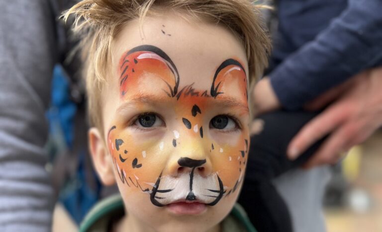 Young child with detailed lion face paint in warm orange and brown tones, looking at the camera while standing at the market, with an adult’s arm gently around them.