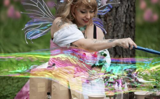 A woman dressed as a woodland fairy creates a large iridescent bubble at an outdoor table covered with craft materials. She wears colourful wings, a flower crown and pastel costume, smiling as the bubble stretches across the scene.