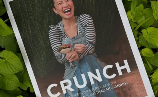 A copy of Poh Ling Yeow’s cookbook “CRUNCH” resting on a bed of fresh green almond leaves. Poh is pictured on the cover, laughing warmly and holding a dish, wearing a striped top and denim apron.