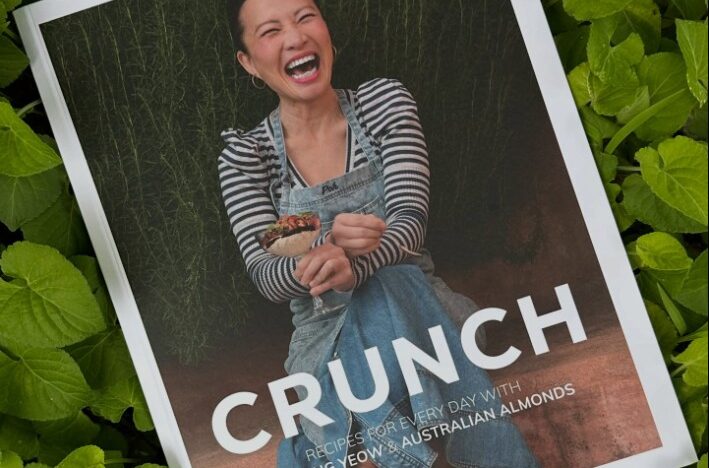 A copy of Poh Ling Yeow’s cookbook “CRUNCH” resting on a bed of fresh green almond leaves. Poh is pictured on the cover, laughing warmly and holding a dish, wearing a striped top and denim apron.