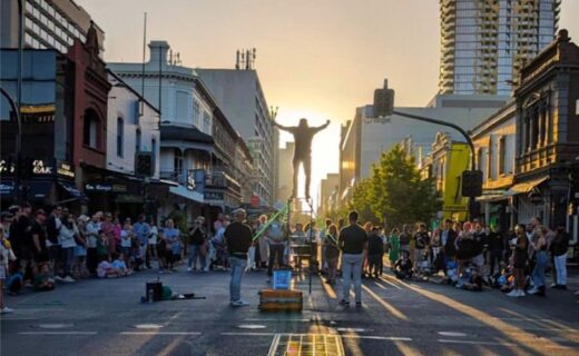 Jesse Uno balancing and juggling on a ladder during a street performance in Adelaide at sunset, surrounded by a large crowd.