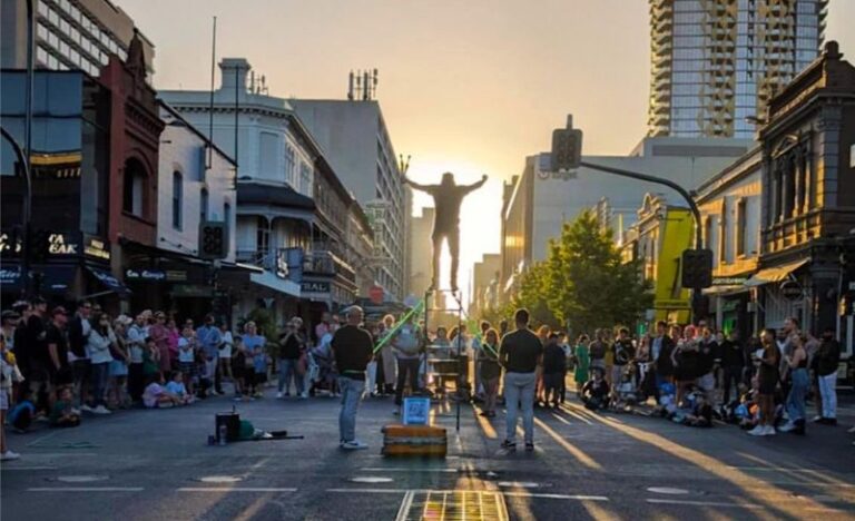 Jesse Uno balancing and juggling on a ladder during a street performance in Adelaide at sunset, surrounded by a large crowd.