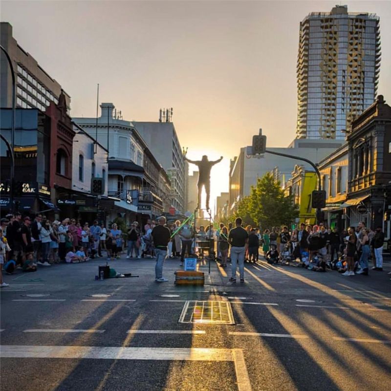 Jesse Uno balancing and juggling on a ladder during a street performance in Adelaide at sunset, surrounded by a large crowd.