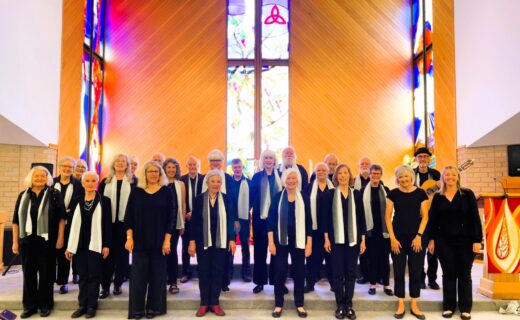 In Good Company choir standing together inside a church with stained glass windows behind them.