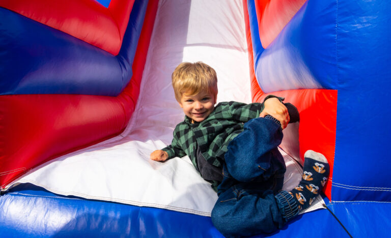 Young child smiling while sitting on a colourful inflatable slide at the farmers’ market.