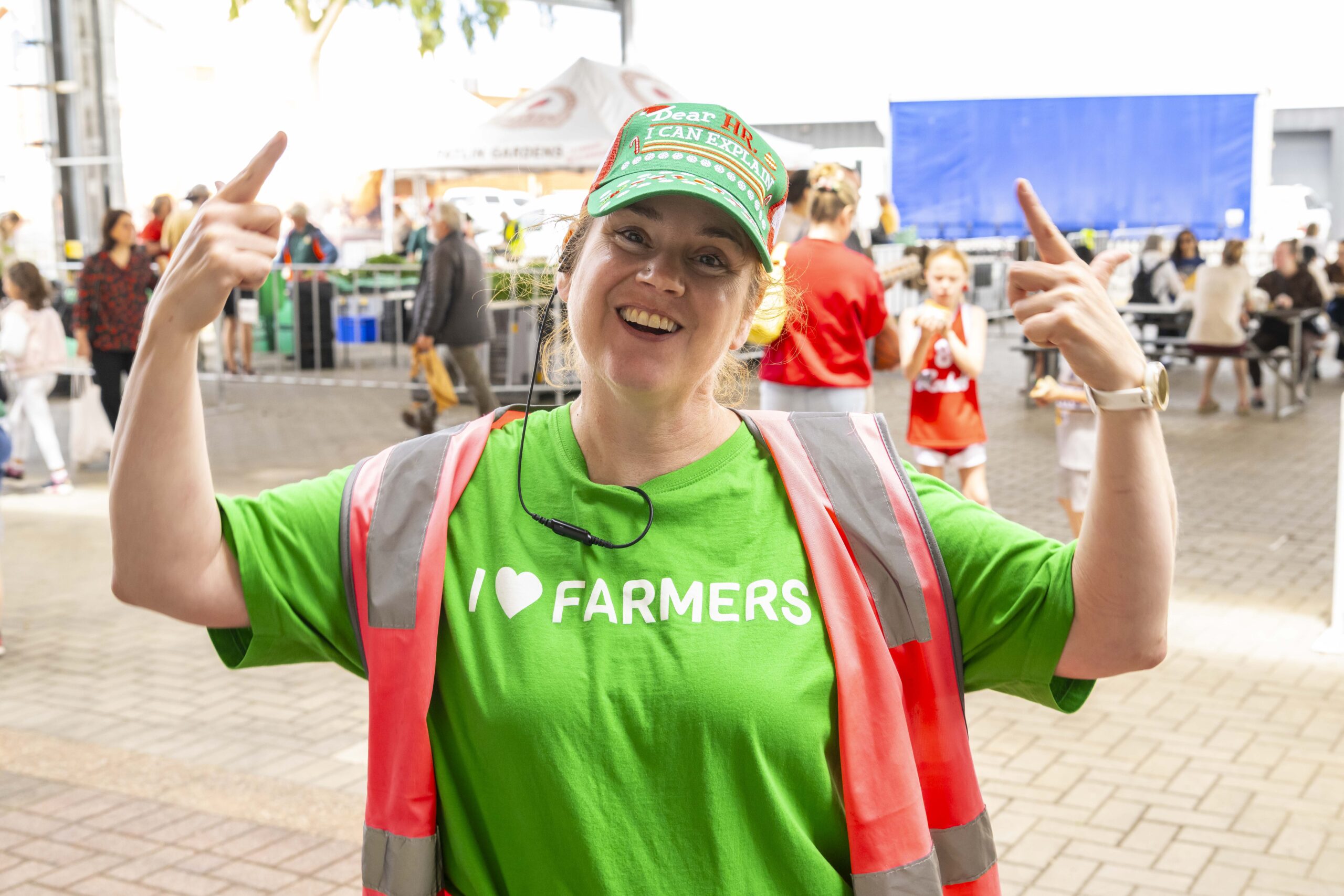 Market volunteer wearing a green “I ♥ Farmers” T-shirt and high-vis vest, smiling and pointing to the message at the Adelaide Showground Farmers’ Market.