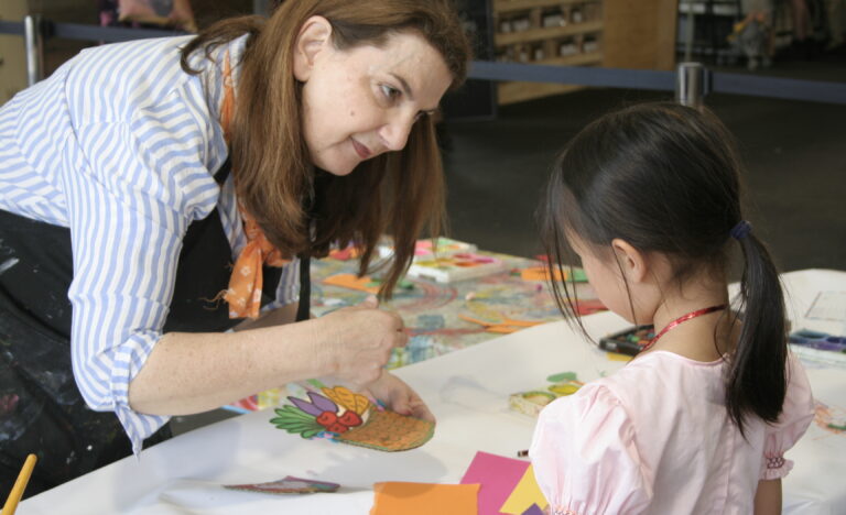 Art facilitator helping a child paint a portrait during a Kids Corner creative workshop inside the market pavilion.