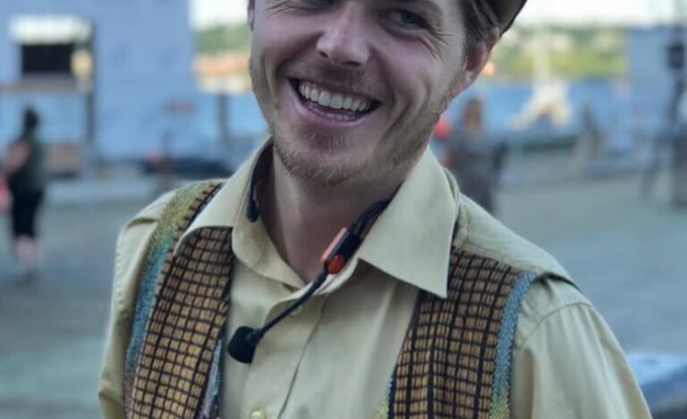 A smiling magician wearing a brown hat and patterned vest stands outdoors, dressed in vintage style attire with a microphone headset, creating a friendly and approachable stage presence.