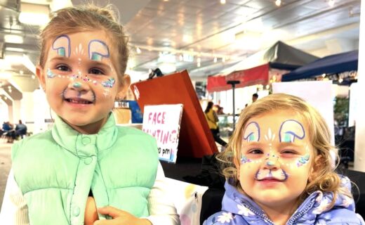 Two young children smiling at the market with colourful bunny face paint, standing indoors near the face painting stall.