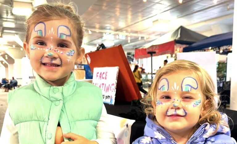 Two young children smiling at the market with colourful bunny face paint, standing indoors near the face painting stall.