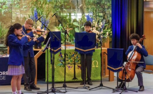 Four junior musicians in school uniforms perform as a string quartet, playing violins, viola and cello on a small indoor stage.
