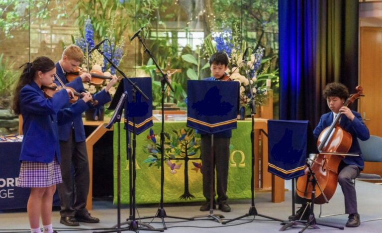 Four junior musicians in school uniforms perform as a string quartet, playing violins, viola and cello on a small indoor stage.