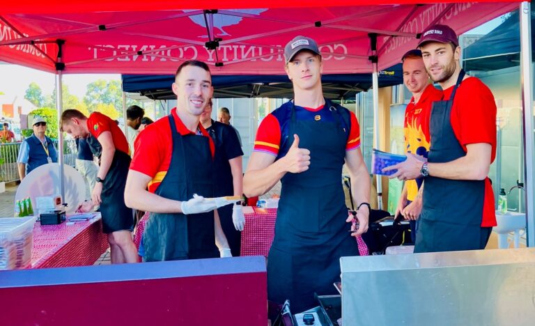 Southern Dragons men’s netball players cooking sausages at the Community Connect stall under a red marquee at Adelaide Showground Farmers’ Market