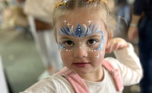 Young child at the market with detailed blue and white face paint design, smiling at the camera.