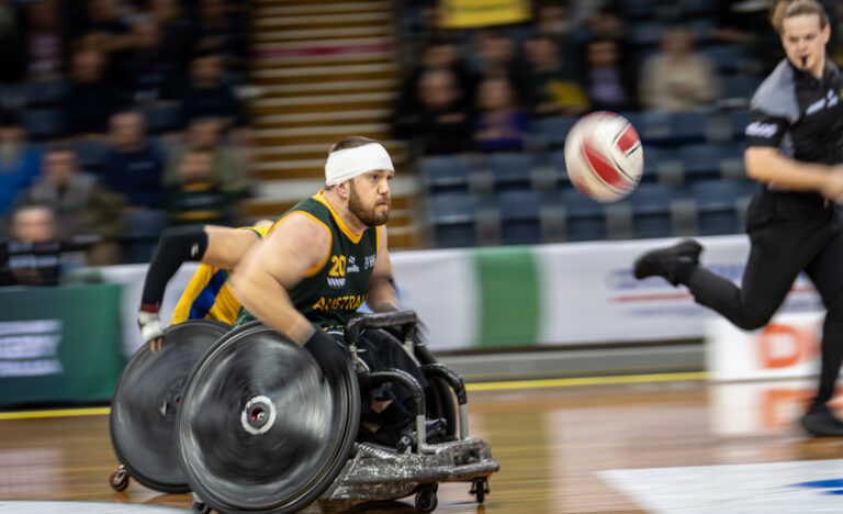 Australian wheelchair rugby player in green and gold jersey racing across court during a fast-paced match, reaching for the ball mid-play
