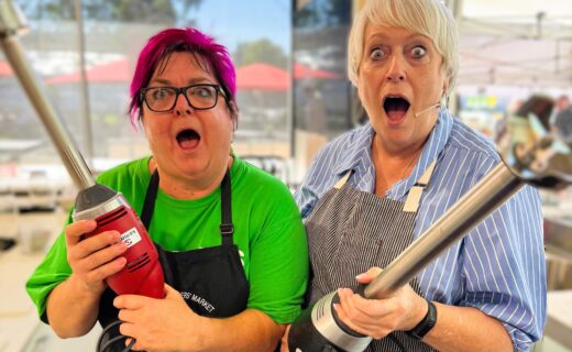 Christine and volunteer Paulie pose in the Market Kitchen holding stick blenders with surprised expressions, ready for another fun cooking demonstration at the Adelaide Showground Farmers’ Market.