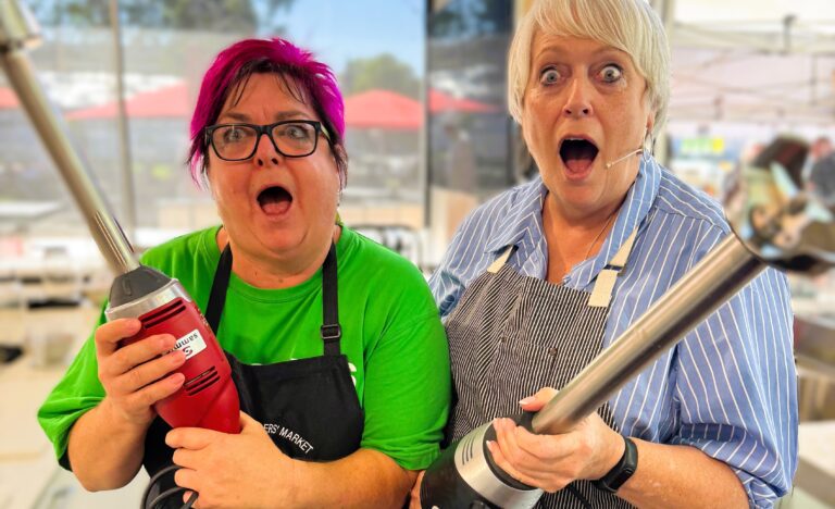 Christine and volunteer Paulie pose in the Market Kitchen holding stick blenders with surprised expressions, ready for another fun cooking demonstration at the Adelaide Showground Farmers’ Market.