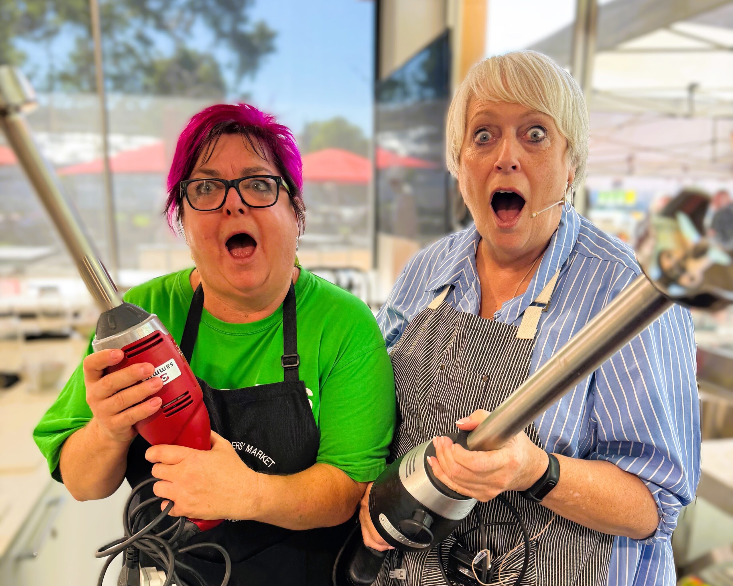 Christine and volunteer Paulie pose in the Market Kitchen holding stick blenders with surprised expressions, ready for another fun cooking demonstration at the Adelaide Showground Farmers’ Market.