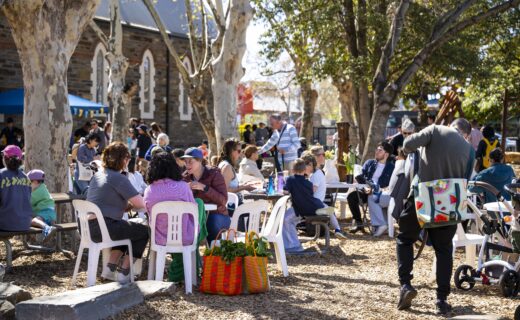 Families and shoppers gather under large shady trees at a community pop up farmers’ market at Goodwood Primary School, enjoying breakfast, coffee and local produce in a relaxed outdoor setting.
