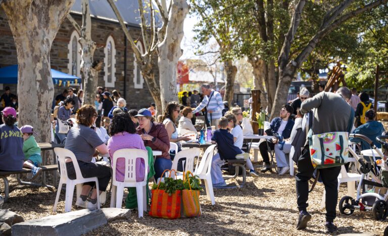 Families and shoppers gather under large shady trees at a community pop up farmers’ market at Goodwood Primary School, enjoying breakfast, coffee and local produce in a relaxed outdoor setting.