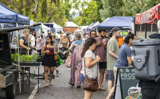Shoppers browse outdoor stalls along Leader Street during a busy Adelaide Showground Farmers’ Market pop up market, with market tents, fresh produce and local food vendors lining the tree filled street.
