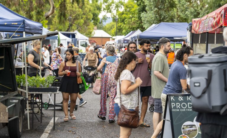 Shoppers browse outdoor stalls along Leader Street during a busy Adelaide Showground Farmers’ Market pop up market, with market tents, fresh produce and local food vendors lining the tree filled street.