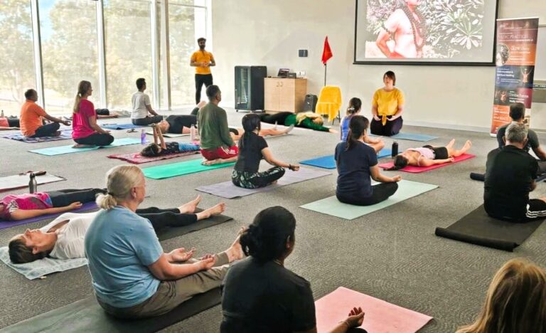 Adults and children taking part in a community yoga and meditation session with Dhyan Foundation Australia, seated on yoga mats indoors during a guided class.