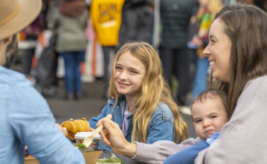 A family enjoying breakfast together at Adelaide Showground Farmers’ Market, with a smiling young girl, mother holding a baby and freshly baked treats on the table.