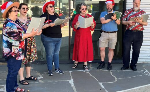 Six members of Cockatoo Chorus singing Christmas carols outdoors, holding songbooks and wearing colourful clothing and festive hats. The Mitchell Park community choir will perform at Adelaide Showground Farmers’ Market on 13 and 20 December 2026.