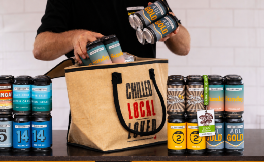 The Suburban Brew craft beer cans being packed into an Adelaide Showground Farmers’ Market insulated jute bag, with a 2027 Market Membership card displayed alongside. The Market will be open on Sunday 27 December 2026.
