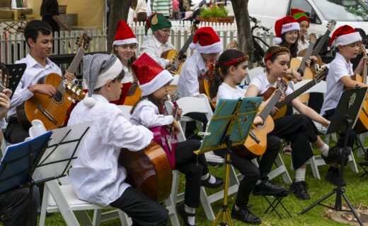 SAYGE musicians wearing Santa hats and festive accessories, seated outdoors playing guitars at the Adelaide Showground Farmers’ Market.