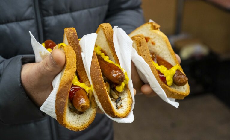 Close up of a market shopper holding three freshly cooked sausages in crusty bread, topped with mustard, tomato sauce and grilled onions.
