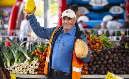 Pat from Patlin Gardens gets into the Gather Round spirit at Adelaide Showground Farmers’ Market, holding up a yellow squash like a footy, surrounded by fresh South Australian vegetables.