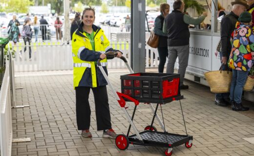 Adelaide Showground Farmers’ Market staff member standing with a red and black V Cart shopping trolley at the Market, showing the prize available to win with a 2027 Membership renewal.